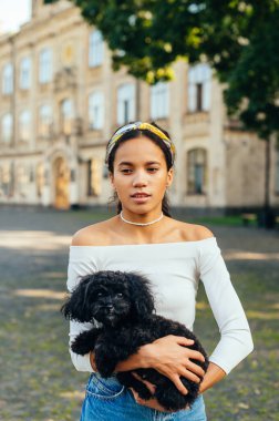 Sıcak bir yaz gününde kollarında sevimli küçük bir köpekle dışarıda dikilen şık gündelik elbiseli neşeli İspanyol kadın. Dikey.