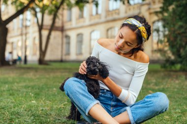 Yazlık giysiler içinde güzel bir kadın çimlerin üzerinde oturur ve sevimli siyah bir köpekle eğlenir. Parkta bir köpekle yürümek.