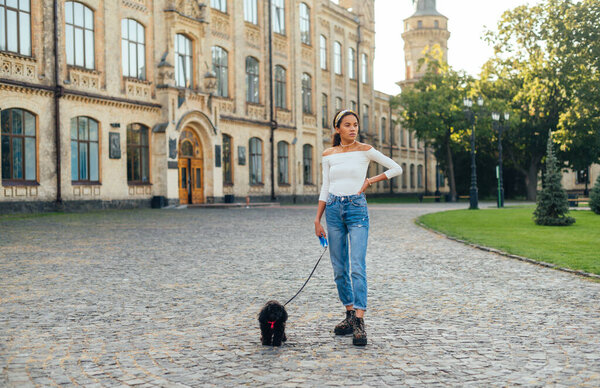 Stylish lady stands on the street of a beautiful old town with a dog on a leash and looks away, full-length photo.