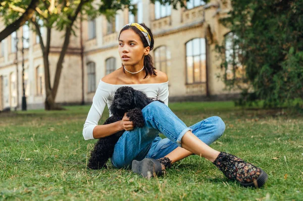 Attractive female owner sitting on the grass with a black little dog breed a toy poodle and looking away with a serious face.