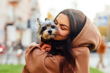 Kapüşonlu siyah saçlı, sevimli Yorkie Terrier 'ine sarılan beyaz bir kadının yakın plan portresi. Bulanık arkaplan.