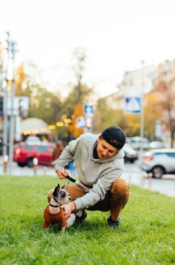 Meşgul bir caddenin önündeki yeşil çimlerde oturan ve sevimli köpeği Yorkshire Teriyeri 'ni okşayan genç bir adamın dikey görüntüsü..
