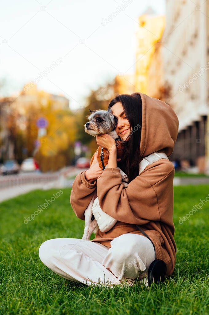 Vertical portrait of a cheerful female dog-owner with her eyes closed in casual outfit, holding her cute yorkshire terrier and sitting on the lawn at the city street.