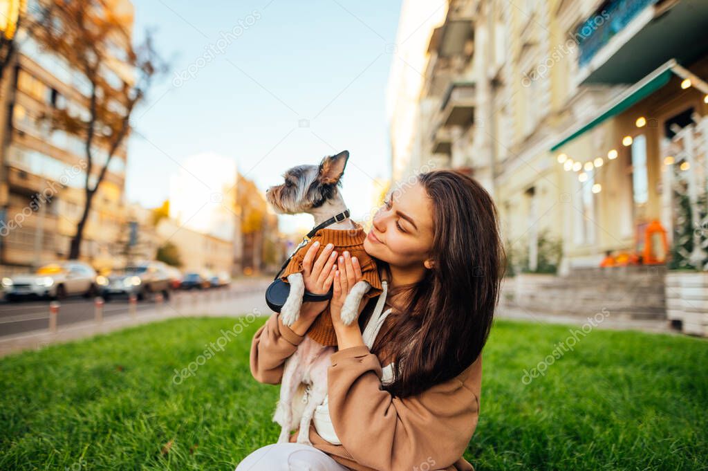 Portrait of a cheerful female dog-owner in casual outfit holding her cute yorkshire terrier and sitting on the lawn at the city street.