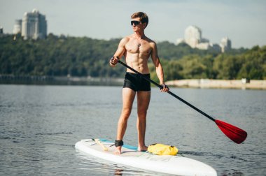 Çıplak gövdeli yakışıklı genç bir adam elinde bir kürekle yemek tahtasının üzerinde duruyor ve nehrin kenarına doğru yüzüyor. Suda aktif eğlence..