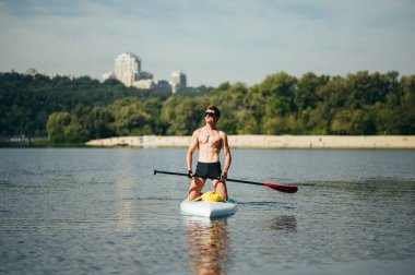 Ellerinde küreği olan atletik bir adam güneşli bir yaz gününde sup tahtasıyla denizde yüzer. Rekreasyon