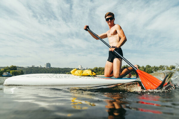 Positive athletic young man surfs on a sup board on a pond, actively paddles on the water and swims fast. Water entertainment on a hot summer day.