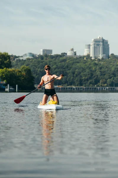 Athletic young man kneels on a sup board and swims on the river against the backdrop of a beautiful landscape. Vertical