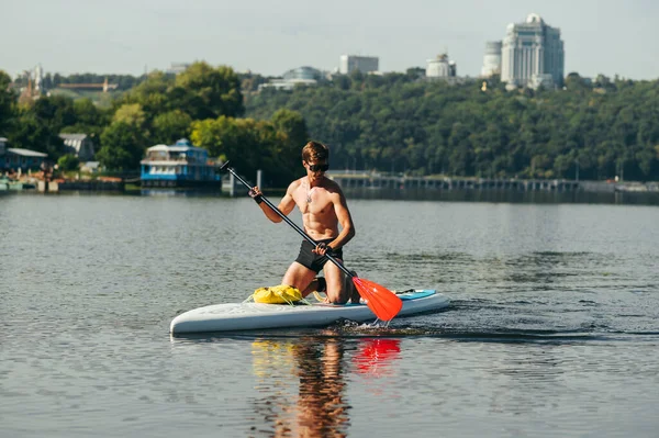 Athletic muscular man in sunglasses swims down the river on a sup board and paddles. Surfing on the river on a sup board