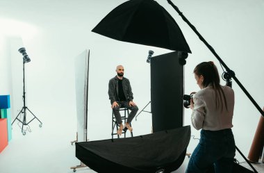 Elinde kamera olan kadın fotoğrafçı, beyaz arka plandaki fotoğraf stüdyosunda sakallı ve yakışıklı bir adam için fotoğraf çekimi yapıyor..