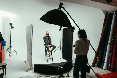 Kadın fotoğrafçı yakışıklı genç bir adam için portre yapıyor, beyaz arka planda bir sandalyeye oturuyor ve yüzünde bir gülümsemeyle poz veriyor..