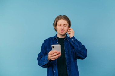 Handsome student guy with smartphone in hand listens to music in wireless headphones with closed eyes isolated on blue background.
