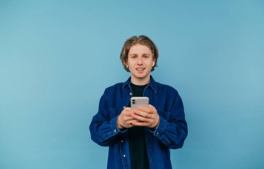 Positive guy in a blue shirt and wireless headphones stands on a blue background with a smartphone in his hands and looks at the camera with a smile on his face.