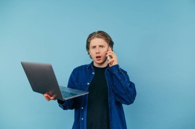 Emotional young man in a shirt talking on the phone and using a laptop on a blue background, looking at the camera and calling with a surprised face.