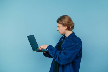 Concentrated young man in a blue shirt stands with a laptop in his hands on a blue background and works with a serious face. Freelancer guy working online, isolated.