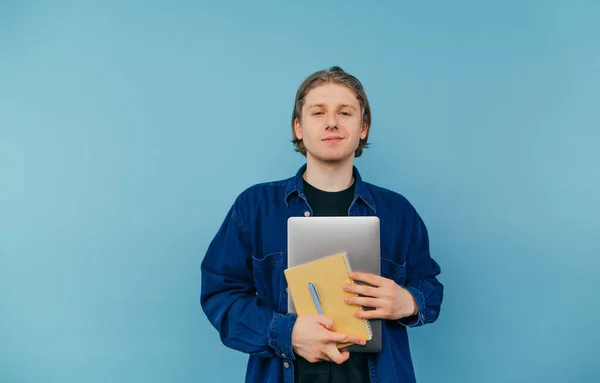 Positive guy in a blue shirt with a laptop and a notebook in his hand stands on a colored background and poses for the camera with a smile on his face. Isolated.
