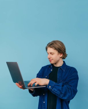 Young male freelancer in roe deer clothes stands with a laptop in his hands on a blue background and works with a smile on his face. Vertical.