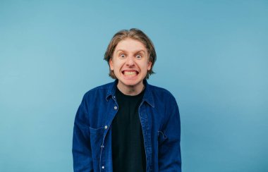Emotional guy in a shirt clenching his teeth looking into the camera with a crazy face isolated on a blue background. Mad young man on a blue background