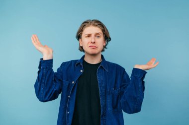 Portrait of a puzzled young man with long hair and in a blue shirt on a blue background, spreads his arms to the sides and looks at the camera.