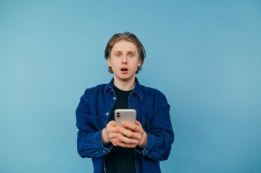 Surprised handsome male student in shirt holding smartphone and looking at camera with shocked face isolated on blue background.