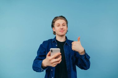 Smiling handsome young man with a long waist and in a blue shirt stands with a smartphone in his hand on a blue background and shows his hand gesture Like, looking at the camera. Isolated.