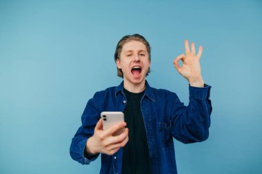 Cheerful young man in casual clothes stands on a blue background with a smartphone in his hands, looks at the camera with a smile on his face and shows an OK gesture.