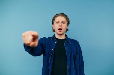 Shocked young man in casual clothes stands on a blue background and points a finger at the comers with an emotional face, wears a blue shirt, isolated.