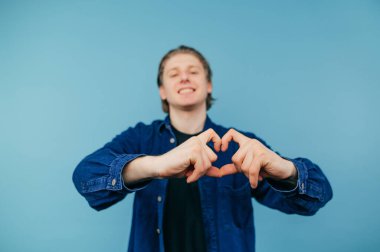 Smiling guy in a blue shirt stands on a colored background, looks at the camera with a happy face and shows a gesture of love. Heart gesture, close up