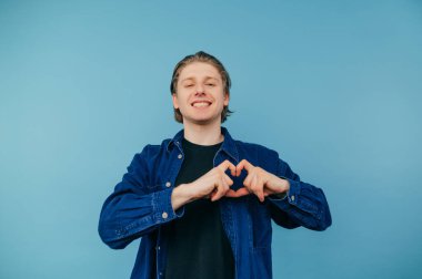 Happy handsome young man in casual clothes and smile on face shows heart gesture to camera, isolated on blue background, wearing blue shirt.