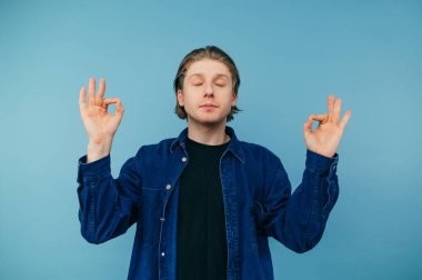 Young man in a shirt and with long hair meditates with his eyes closed on a blue background. Guy student doing yoga, isolated.