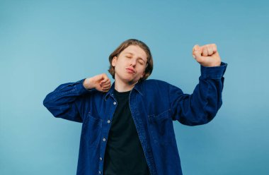 Mavi arka planda gözleri kapalı dans eden tişörtlü komik öğrenci. Genç adam renkli bir arka planda dans ederken rahatlıyor. İzole edilmiş.