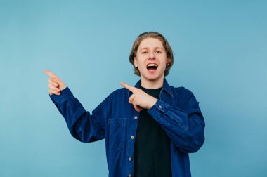 Sıradan giyinmiş neşeli yakışıklı genç adam mavi bir arka planda duruyor yüzünde bir gülümsemeyle kameraya bakıyor ve parmaklarını boş bir koltuğa doğrultuyor, mavi bir gömlek giyiyor..