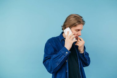 Uzun saçlı ve gömlekli yakışıklı genç adam mavi arka planda duruyor ve ciddi bir yüzle akıllı telefondan iletişim kuruyor.