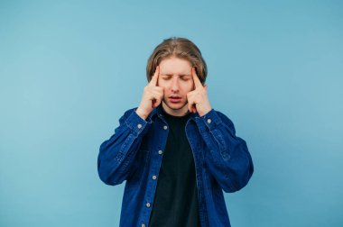 Sıradan giyinmiş bir adam mavi arka planda durur ve baş ağrısından parmaklarını şakağına geçirir, gözlerini kapatır ve acı çeker..