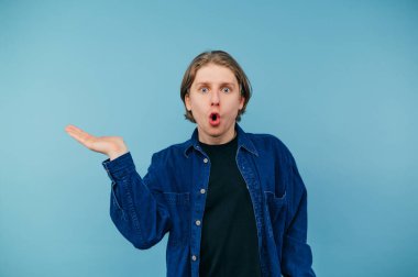 Şok olmuş gömlekli adam mavi arka planda duruyor, duygusal olarak kameraya bakıyor ağzı açık ve elinde bir fotokopi odası tutuyor. İzole edilmiş.