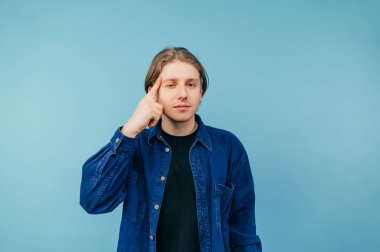 Tişörtlü neşeli adam mavi arka planda duruyor ve işaret parmağını şakağında gösteriyor ve yüzünde hafif bir gülümsemeyle kameraya bakıyor..