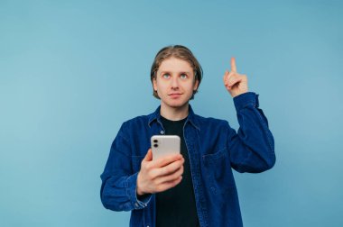 Gömlekli yakışıklı bir adam elinde akıllı bir telefonla mavi bir arka planda duruyor ve yüzünde bir gülümsemeyle parmağını boş bir yere doğrultuyor..