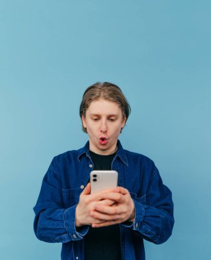 Tişörtlü şaşkın adam elinde akıllı bir telefon tutuyor ve mavi arka planda izole edilmiş şok olmuş bir yüzle ekrana bakıyor..