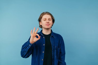 Mavi arka plandaki olumlu genç adam parmaklarıyla ve yüzünde bir gülümsemeyle mavi bir gömlek giydiğini gösteriyor..