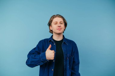 Tişörtlü neşeli öğrenci mavi arka planda duruyor ve yüzünde gülücükler, izole bir şekilde baş parmaklarını gösteriyor..