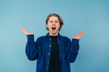 Sıradan giyinmiş neşeli bir adam ve şaşırmış bir yüzle kameraya baktı ve mavi bir arka planda tek başına ellerini kaldırdı..