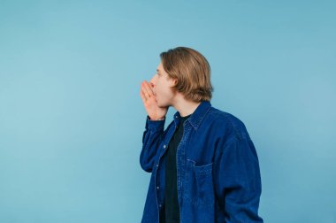 Mavi gömlekli, uzun saçlı, leğen kemikli genç adam avucunu mavi bir cephede gizliyor. Boşluğu kopyala