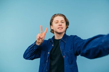 Uzun saçlı yakışıklı genç adam mavi arka planda selfie çekiyor ve yüzünde bir gülümsemeyle barış jesti sergiliyor..