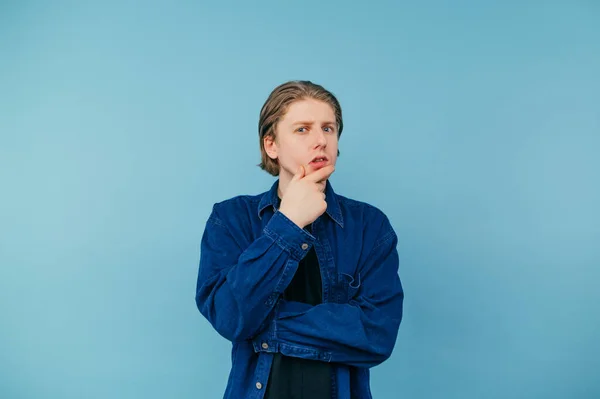 Pensive guy student in shirt isolated on blue background, looking at camera with pensive face.