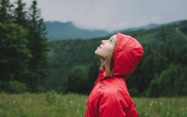 Kırmızı yağmurluklu çekici turist kız portresi dağlardaki vadide duruyor ve bulutlu gökyüzüne bakıyor. Yağmurluk giymiş güzel bir kadın yağmur bulutlarına bakıyor..