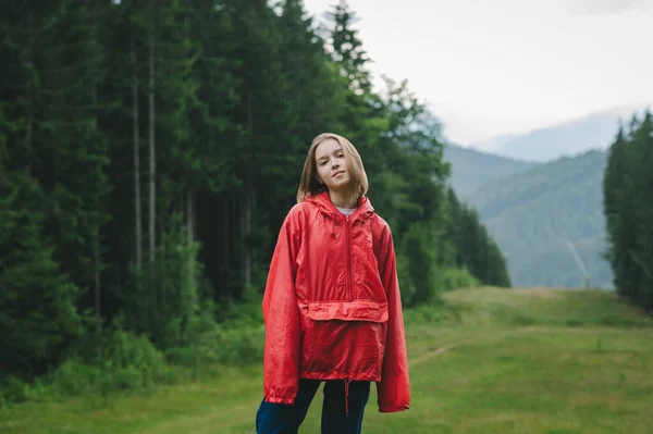 Attractive hiker girl in a red raincoat rainbow stands on a mountain in the rain against a background of mountain forests and fog, looks into the camera and smiles. Hiking in the mountains in the rain