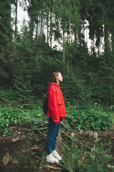Hiker girl stands in forest in red raincoat and looks away. Attractive girl hiking in the forest, stands on a trunk on a mountain stream. Vertical