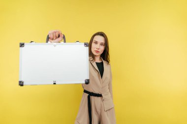 Ciddi bir iş kadını ciddi bir yüzle sarı arka planda duruyor ve kameraya metal bir bavul gösteriyor. Bir çanta dolusu parası olan, izole edilmiş bir kadın..