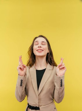 Takım elbiseli tatlı bir kadın, gözleri kapalı bir dilek diler, parmaklarını ve hayallerini, yüzünde bir gülümsemeyle, sarı bir arka planda izole bir şekilde..
