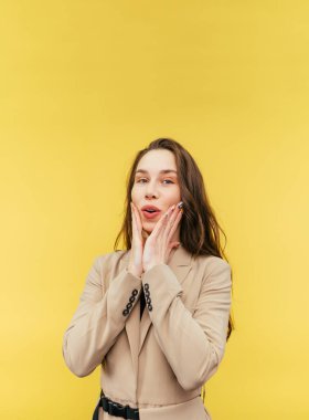 Sarı arka planda izole edilmiş şirin bir bayan yüzünde sevimli bir gülümsemeyle kameraya bakıyor ve kendini yanaklarından tutuyor. Dikey.